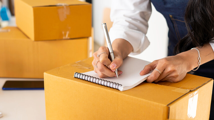 Image of a man writing a list with a pen on a notepad on top of a cardboard box.
