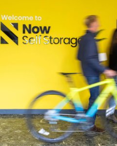 Image of yellow wall with Now Storage logo with a man walking by holding a bicycle.