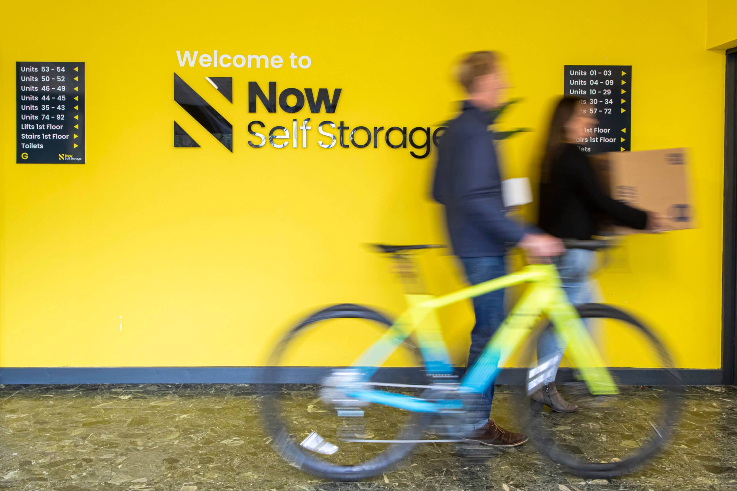 Image of yellow wall with Now Storage logo with a man walking by holding a bicycle.