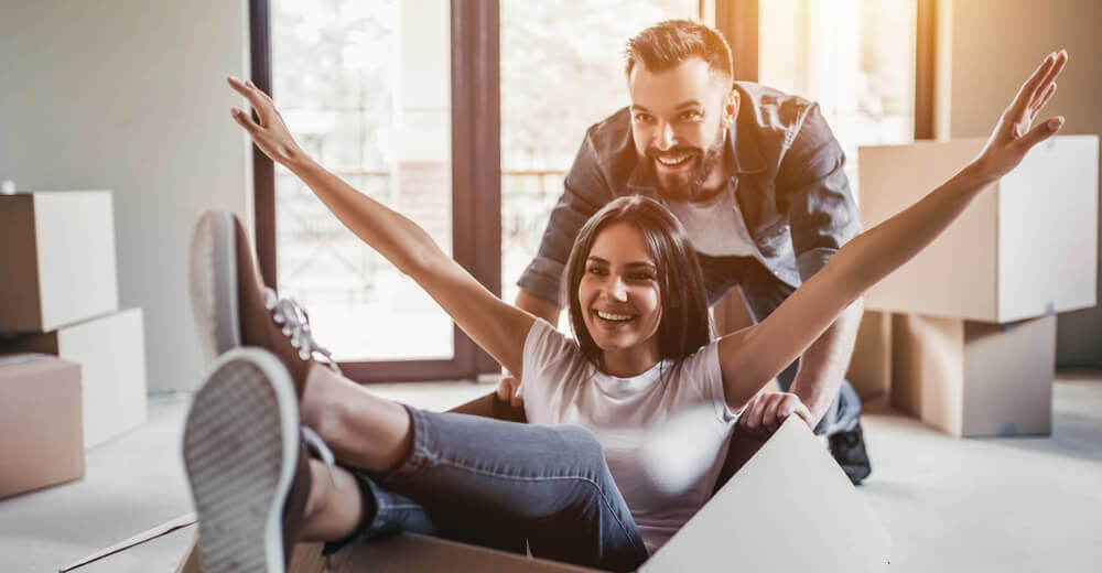 Image of a man and a woman happy while the woman sits laughing in empty cardboard storage box in new home.