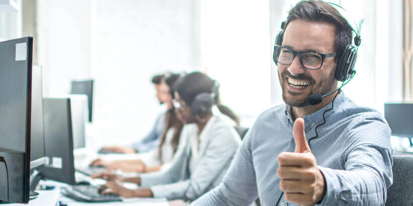 Image of a smiling man with glasses wearing a headset putting his thumbs up while working on office desk in front of computer screen.