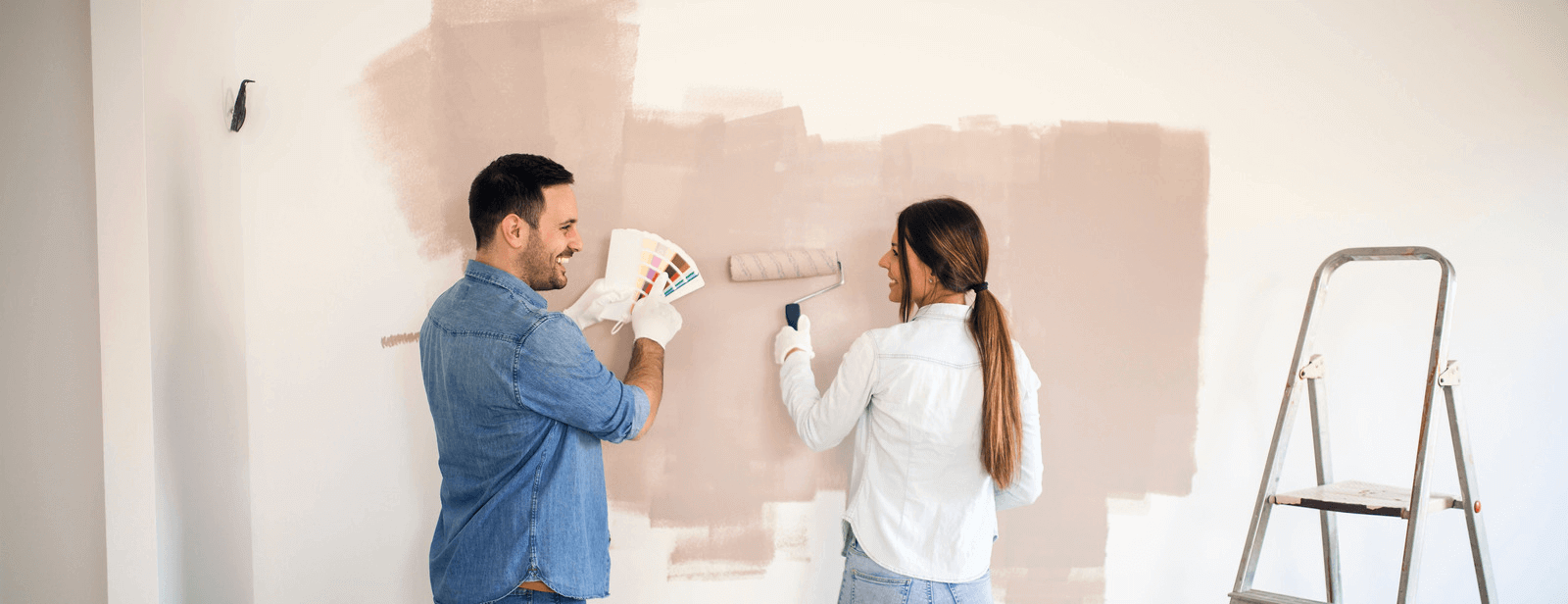 A couple painting their white walls a nude pink shade with rollers.