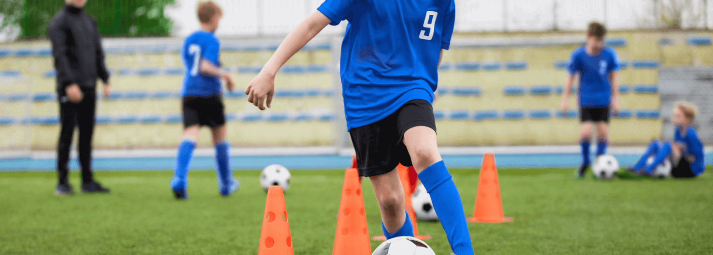 A young boy dribbling the football between cones at football practice.