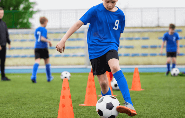A young boy dribbling the football between cones at football practice.