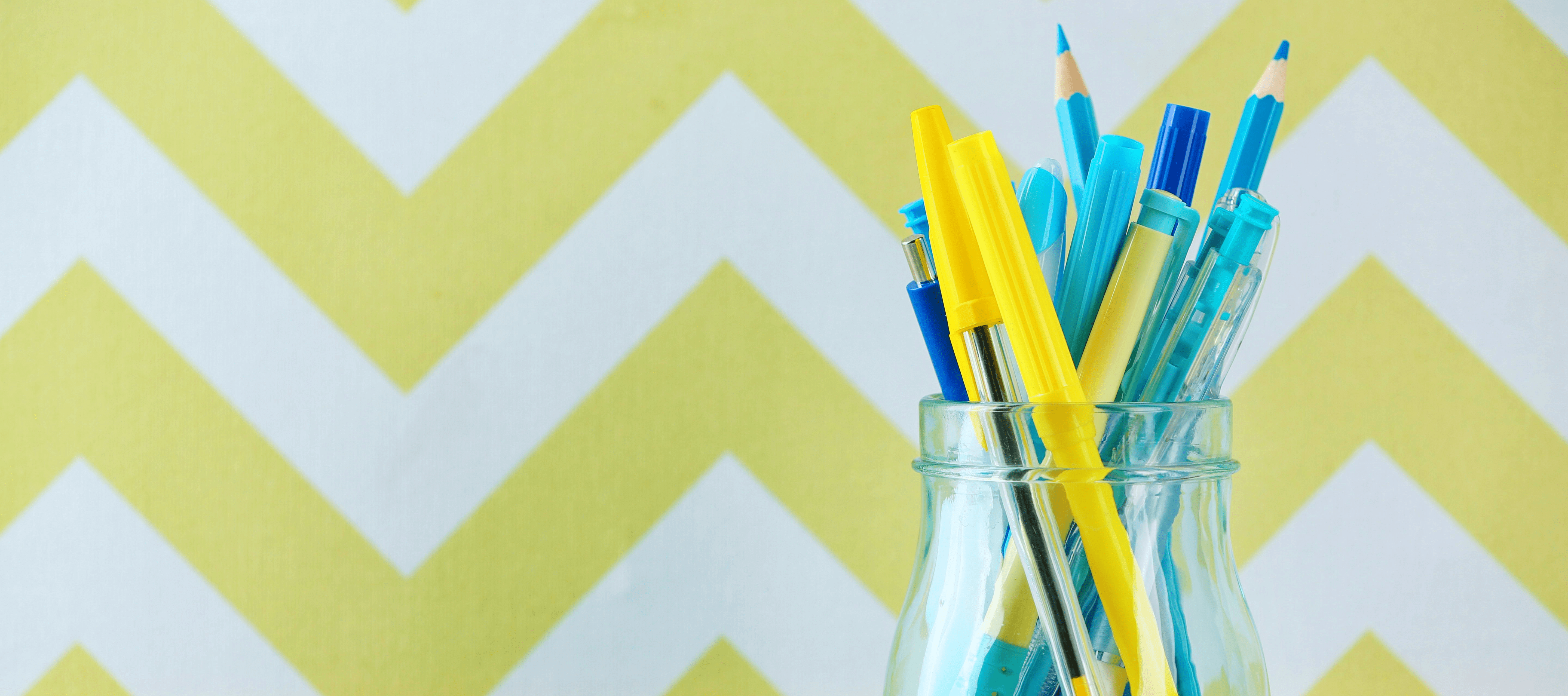 Jar being reused to hold pens and pencils, against a yellow and white herringbone wall pattern.