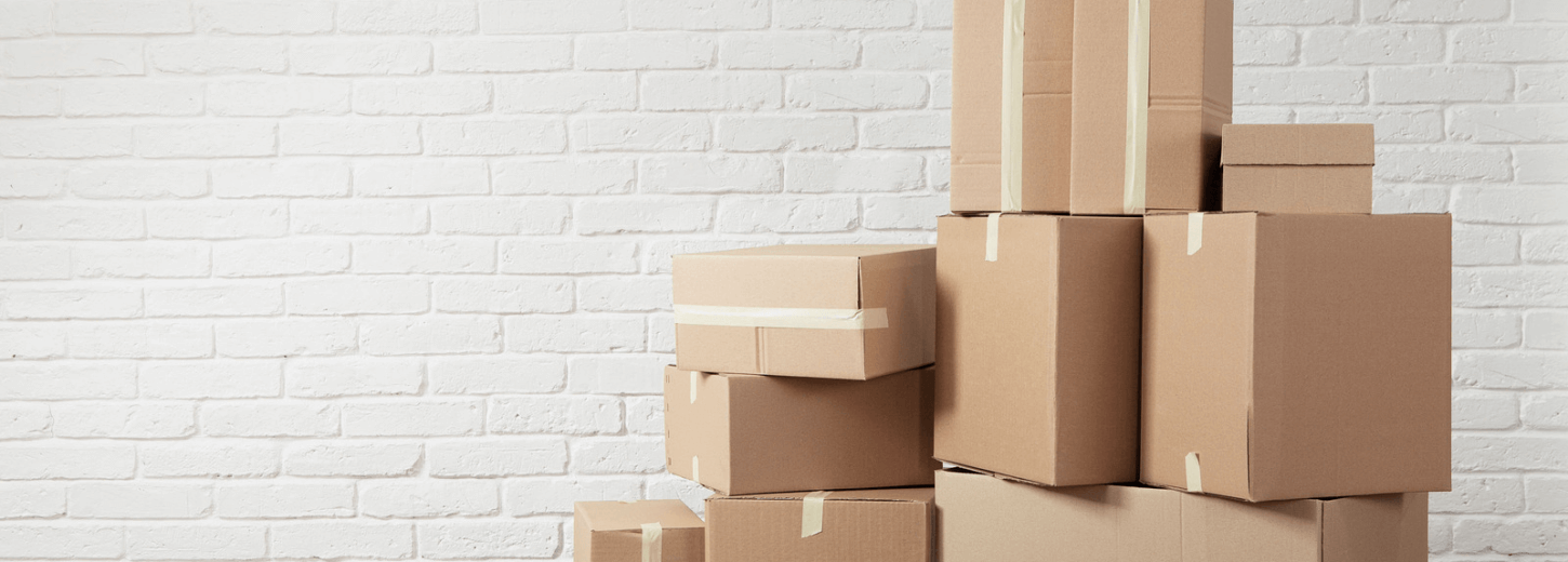 Cardboard boxes piled up ready for moving house against a white brick wall.