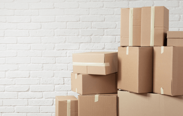 Cardboard boxes piled up ready for moving house against a white brick wall.