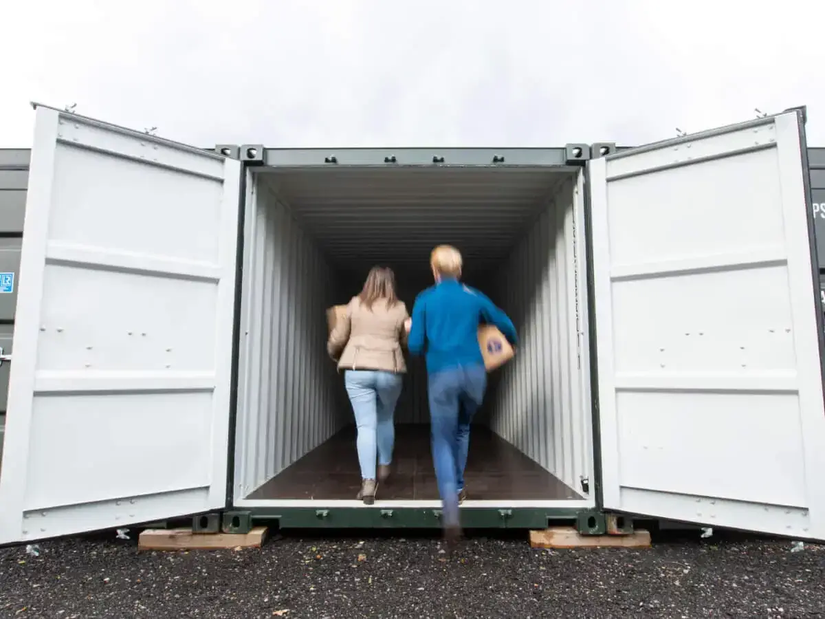 A man and woman carrying boxes into a green storage container
