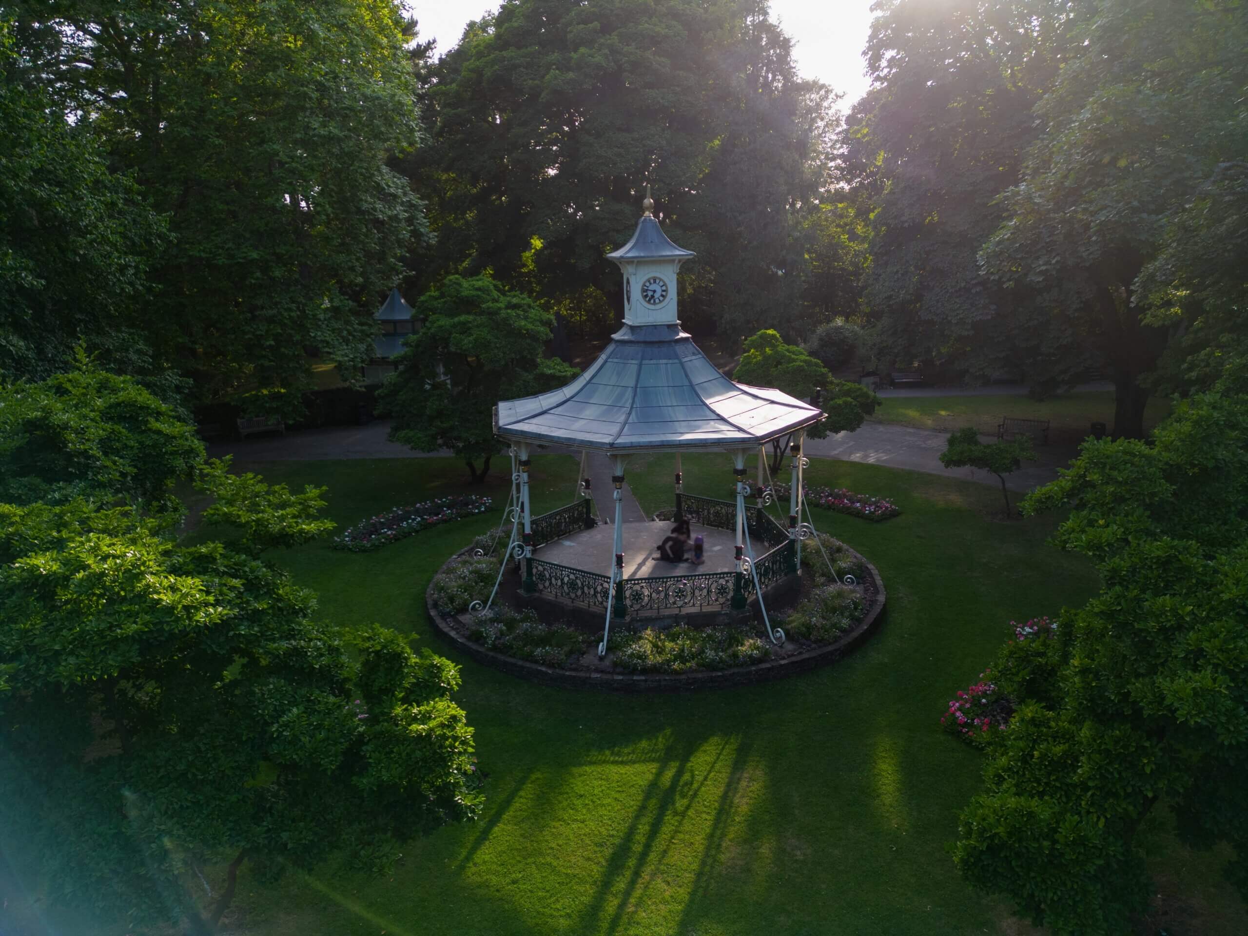 An image of a beautiful English garden and historic bandstand with clock in Swindon Town Gardens at sunset with sun glare.